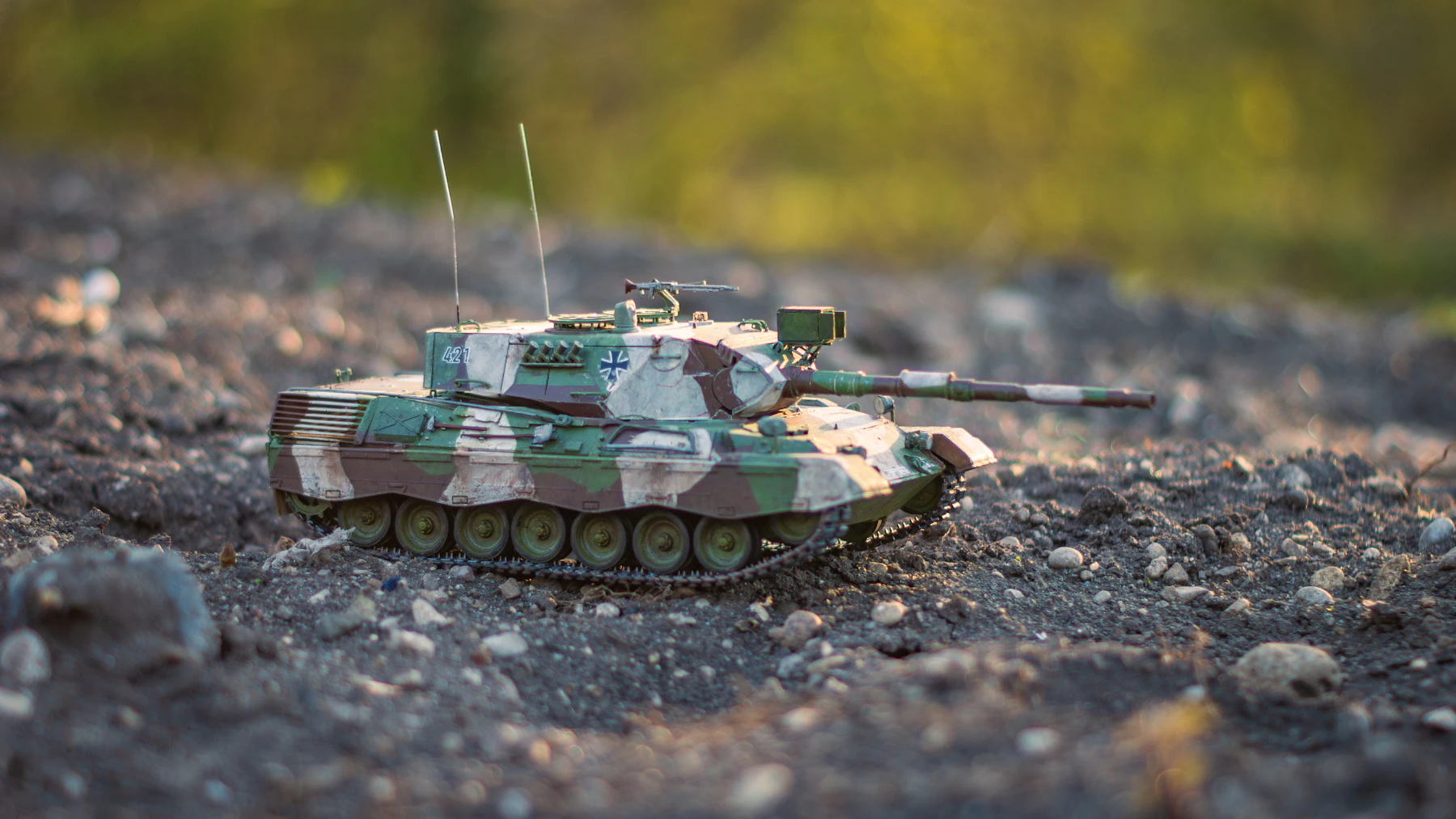 A green, white and brown camouflaged model tank with two antennas crossing dark gravel