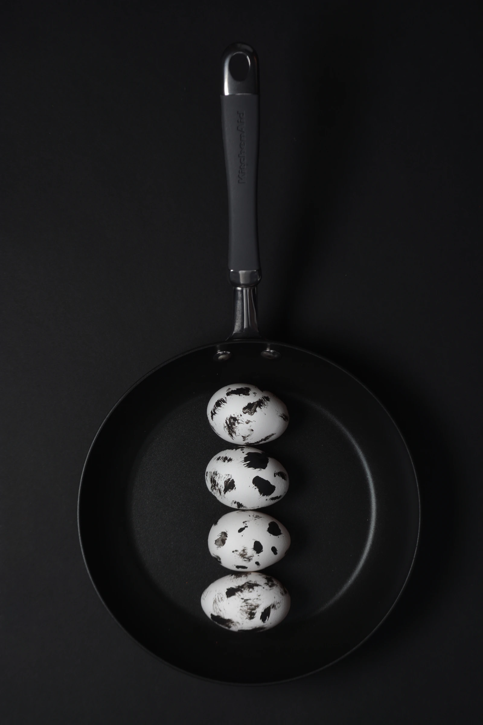 Top-down black frying pan with four speckled quail eggs on a black ground