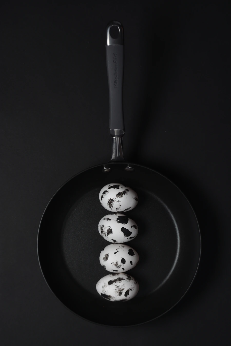 Top-down black frying pan with four speckled quail eggs on a black ground