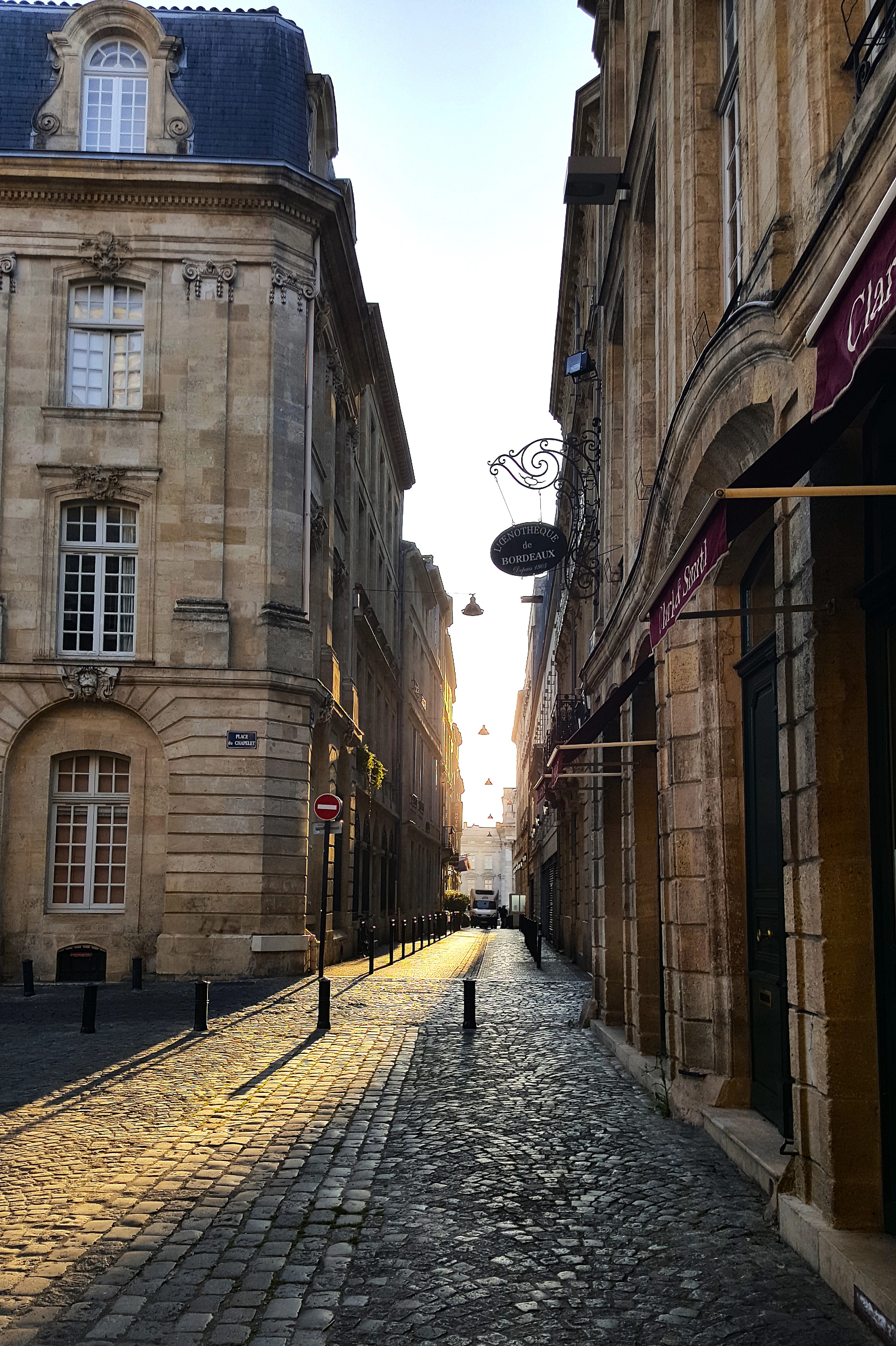 Low evening sun rakes down a narrow cobbled Bordeaux side street, lighting the pale limestone facades and a wrought-iron shop sign hanging out over the empty pavement.