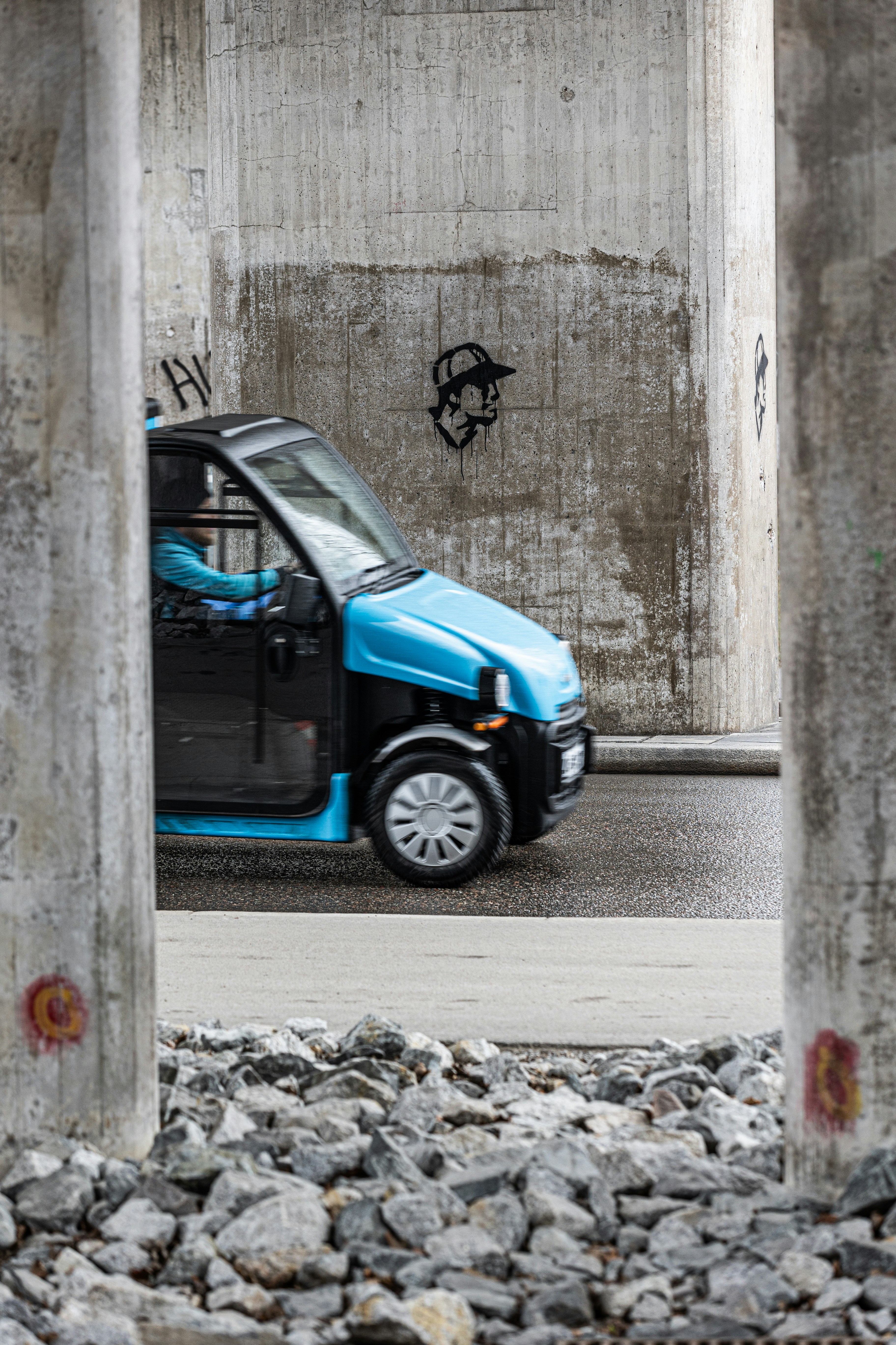 A small blue car drives under a concrete structure.
