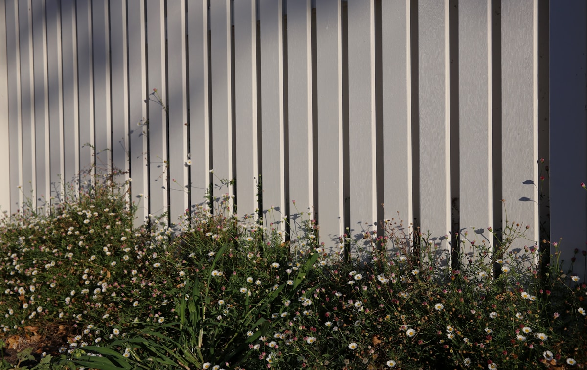 White vertical privacy fence panel with small wildflowers and green grass growing at its base against a grey sky
