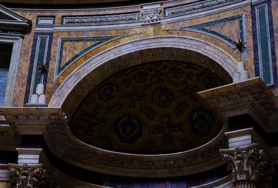 The image shows a close-up view of an ornate archway, likely found within a historic building such as the Vatican Museums in Rome, Italy. The archway is adorned with intricate carvings and decorations, showcasing the craftsmanship of the era it was built. The ceiling above the archway features a large design that adds to its grandeur. Two small statues are perched atop the archway, adding an element of historical significance to the scene.

The image is taken from a low angle, emphasizing the height and majesty of the archway. The colors in the image are predominantly browns and greens, reflecting the natural hues found within historic buildings.