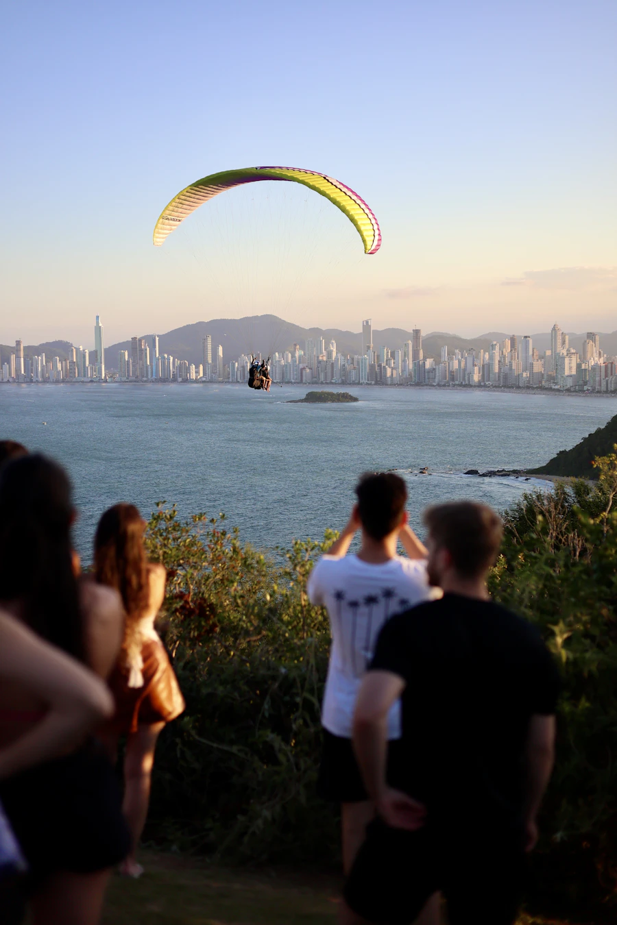Pessoas observando parapente sobre o mar e a cidade
