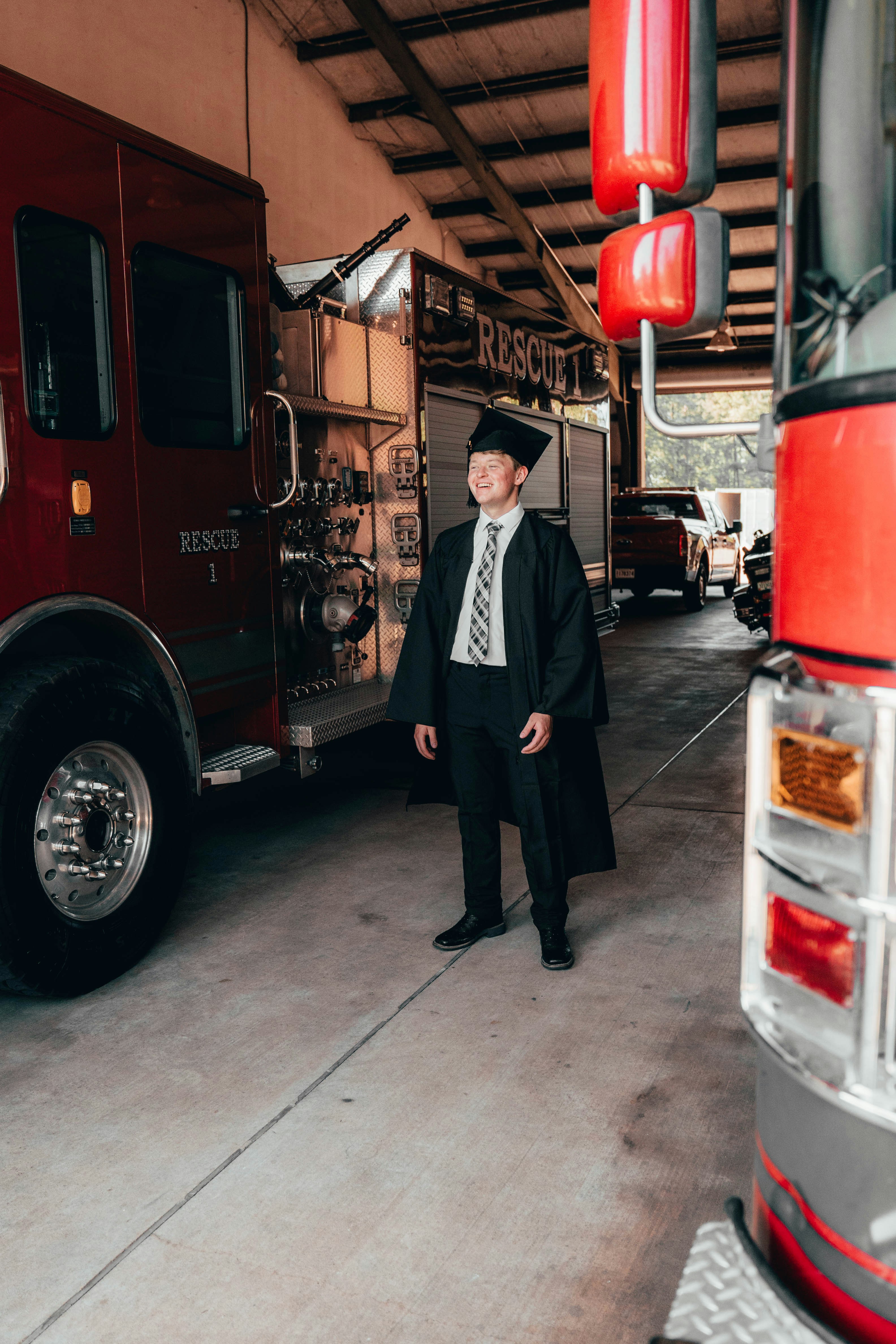 Graduating student in a cap and gown near firetrucks.