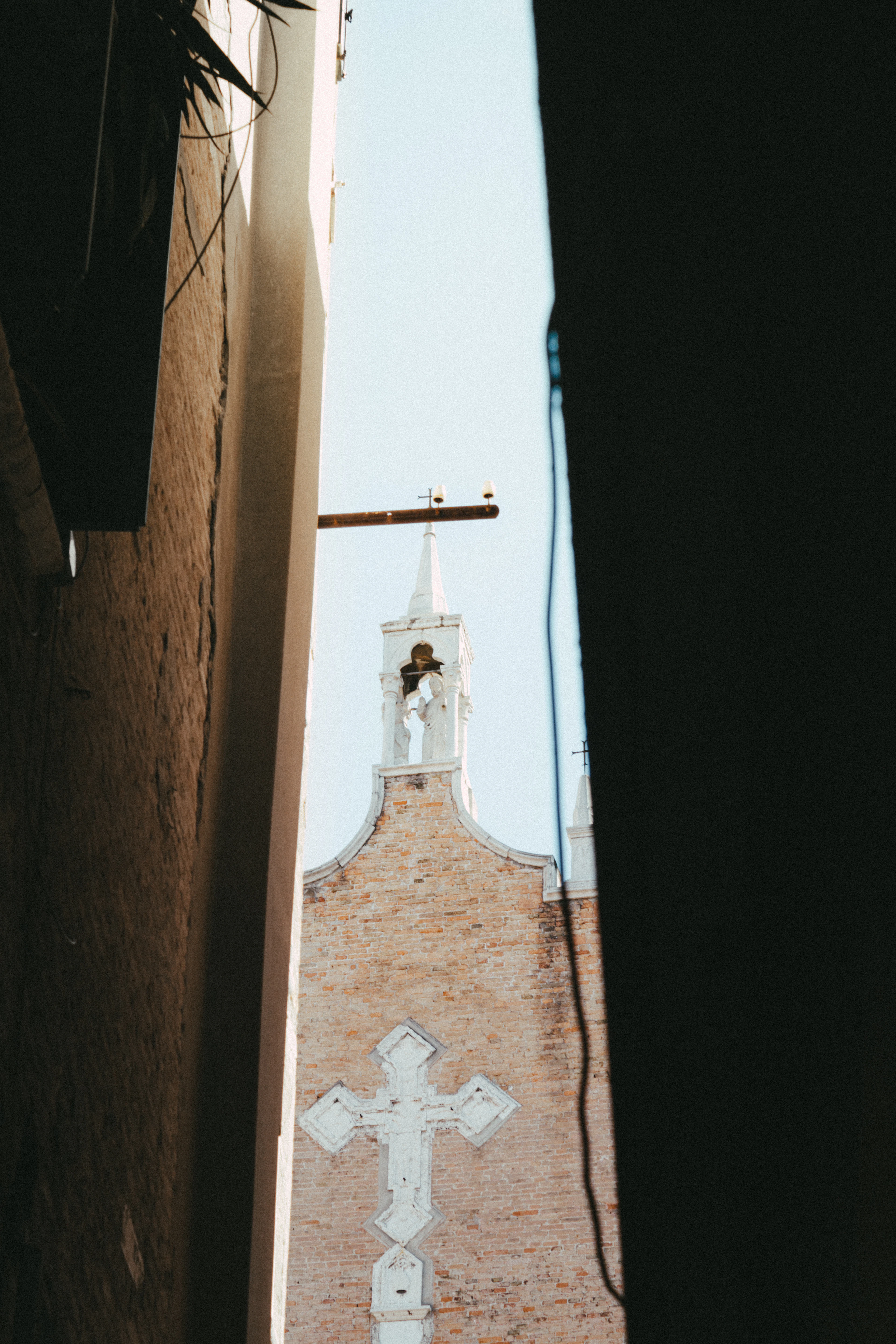 Framed between two dark building walls, a sunlit brick church facade with a cross and bell tower is glimpsed down a narrow alley.