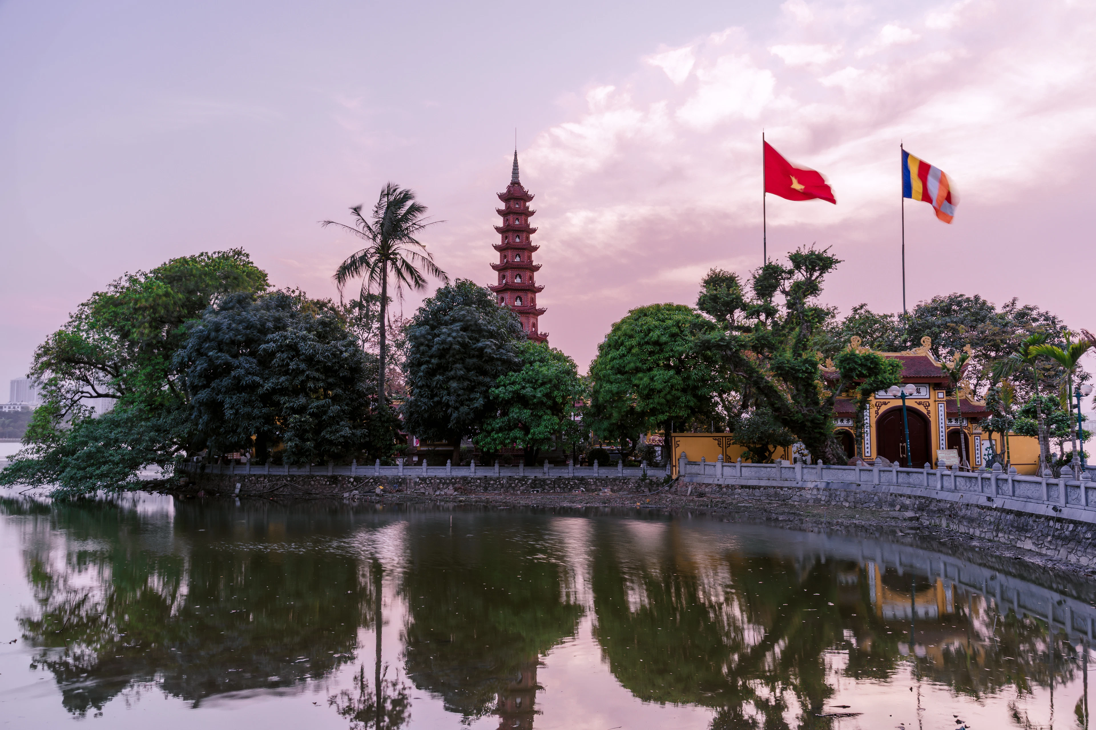 Tran Quoc Pagoda in Hanoi reflected in still water at dusk, flags flying at the gate