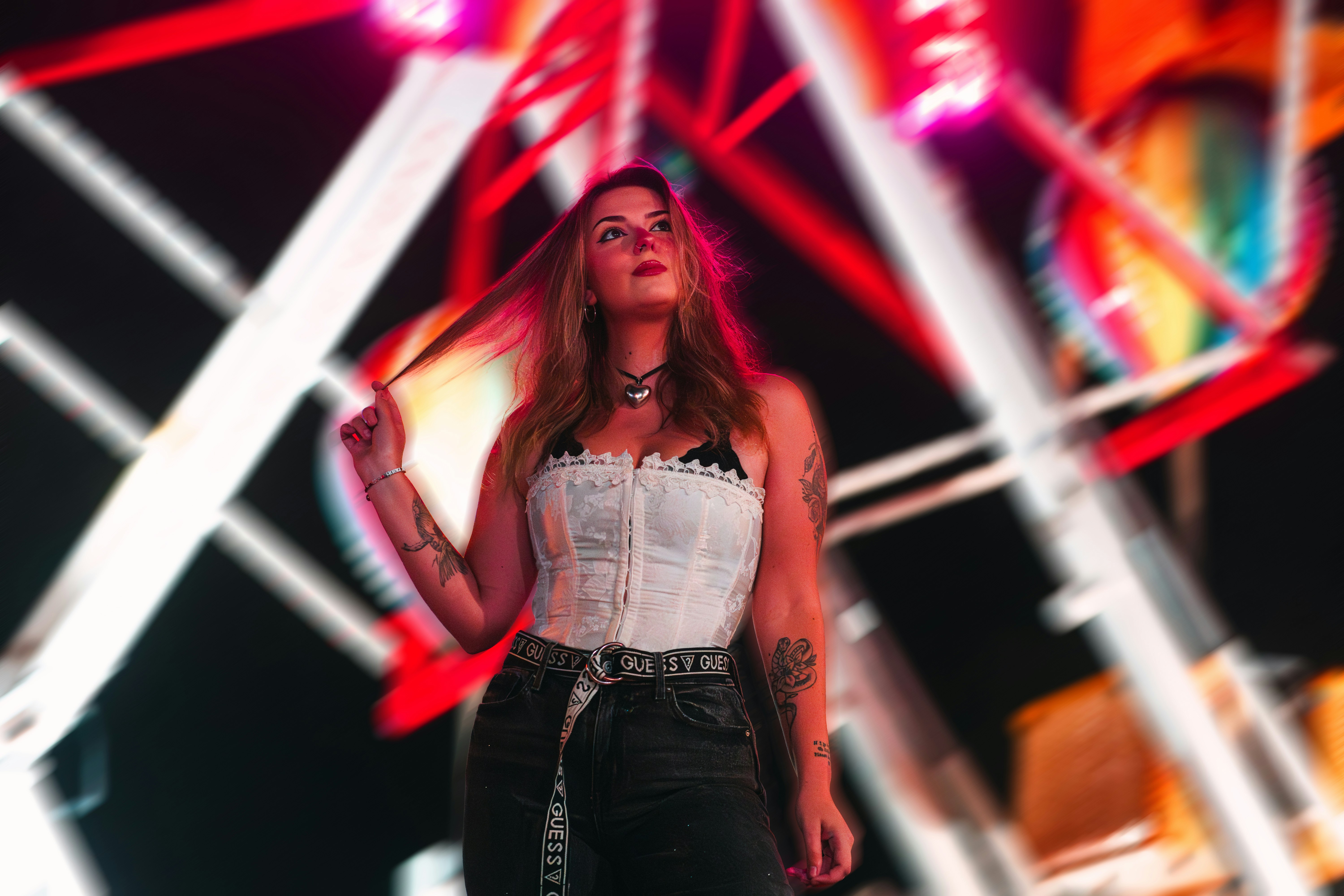 A confident woman stands under the glow of a Ferris wheel at night, lit by vibrant neon and surrounded by the energy of the carnival. Her pose, outfit, and expression channel a fearless sense of self, blending street style with fairground fantasy. A striking portrait that captures the electric mood of nightlife and self-expression.