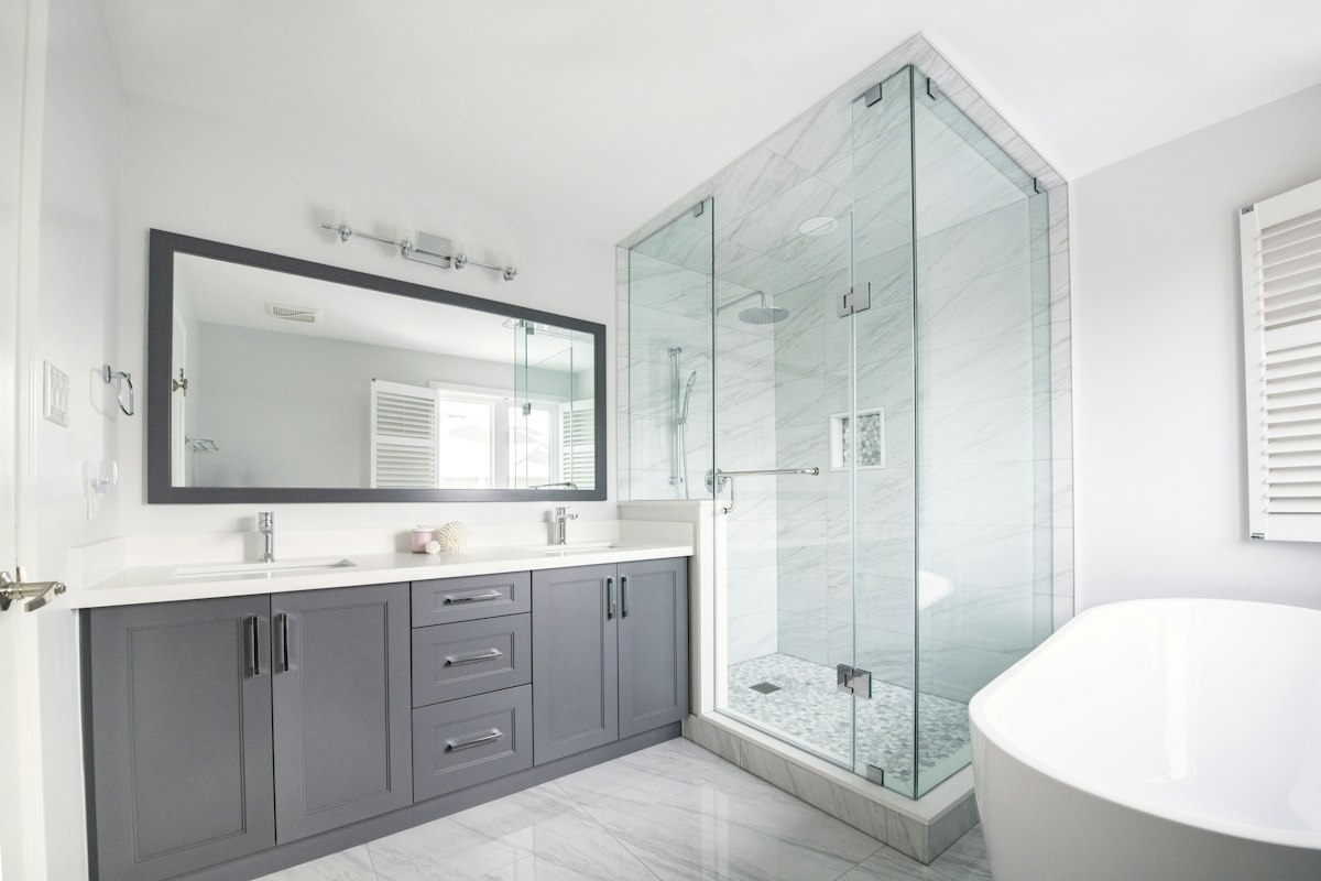 Baño recién reformado con mueble doble de vanitory gris, espejo grande, ducha cerrada con mampara de vidrio y piso de porcelanato blanco veteado, y una bañera exenta en primer plano