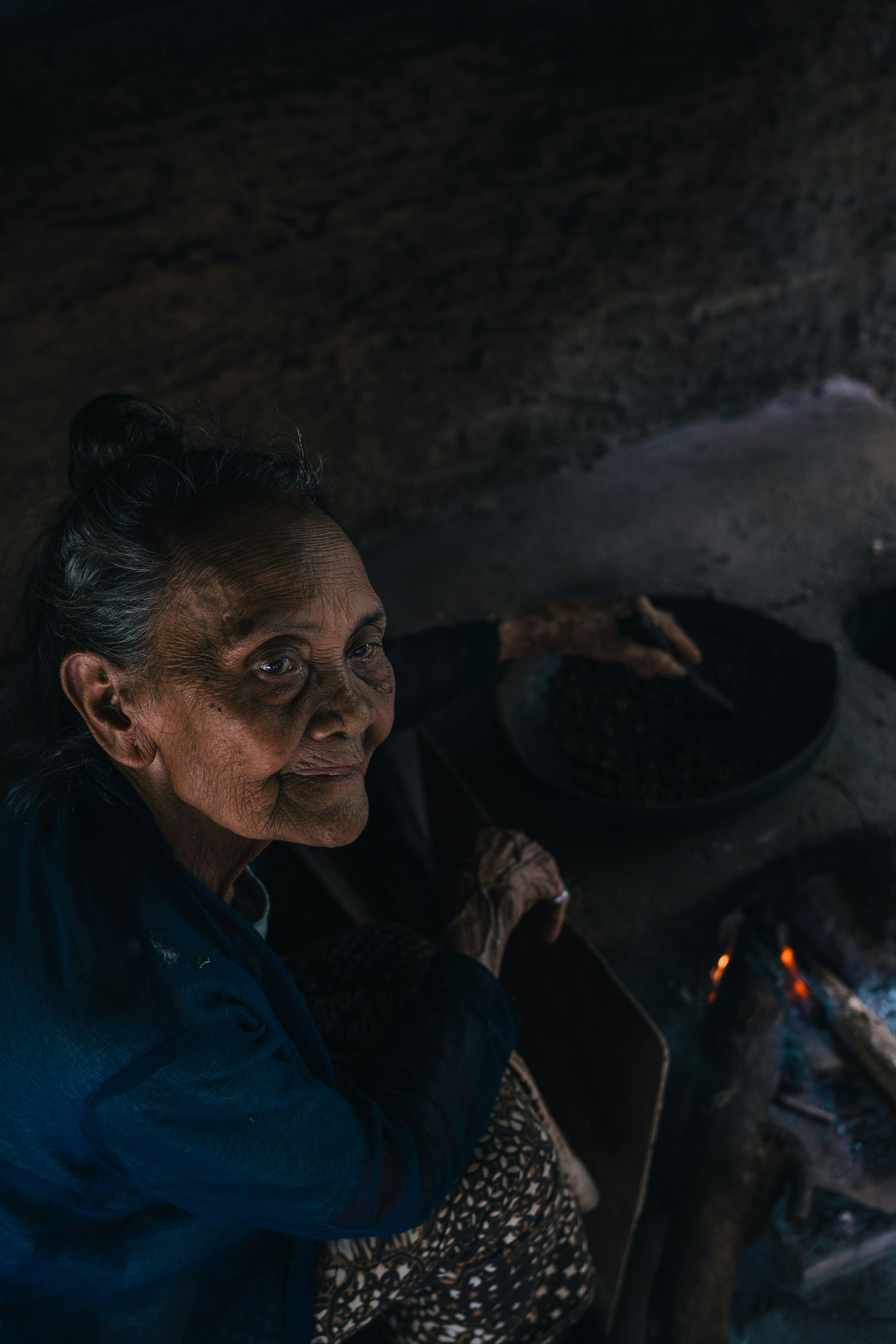 An elderly woman roasts coffee beans over fire.