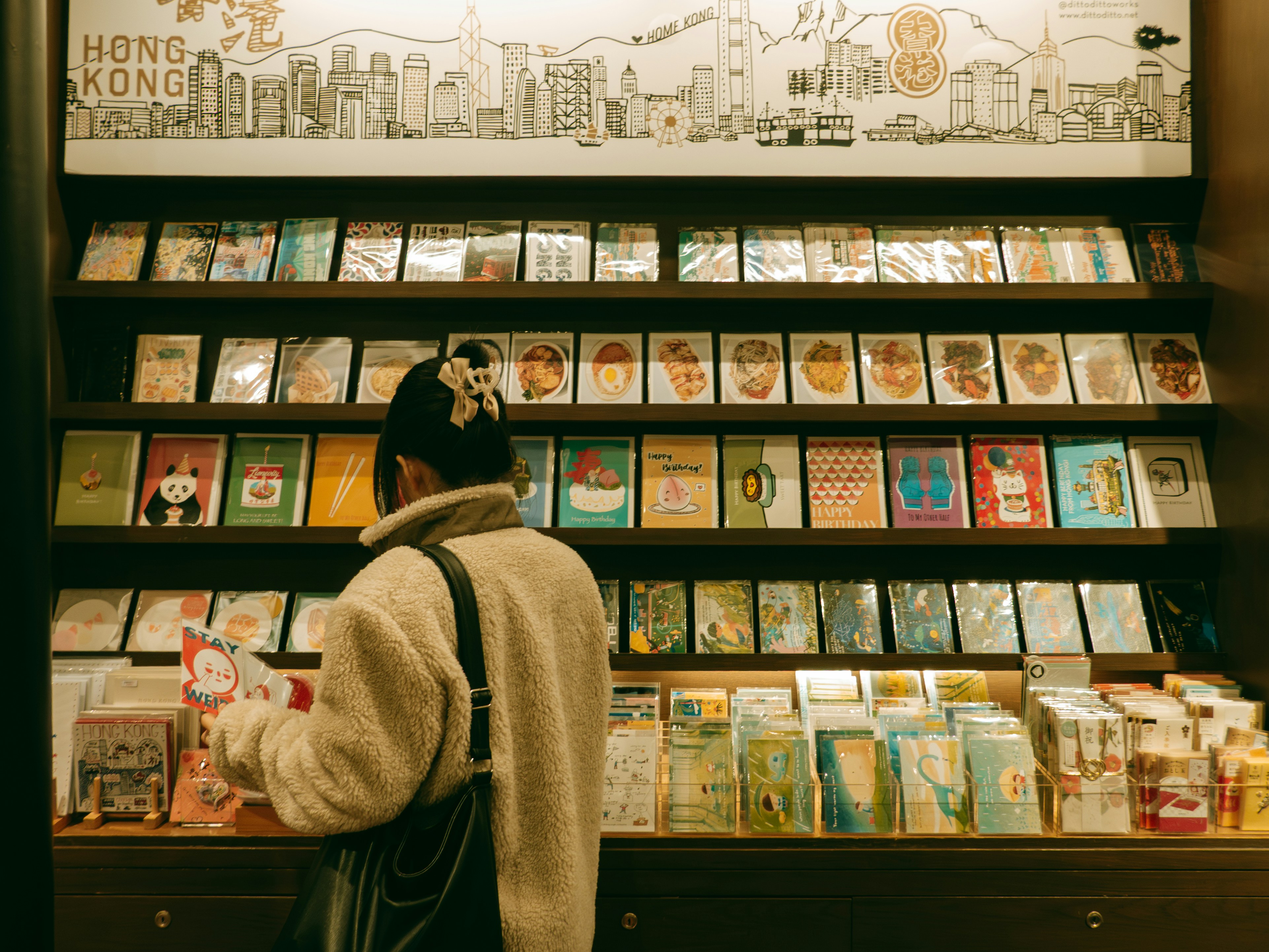 A woman browses postcards in a gift shop., Inside a bookstore in Causeway Bay, Hong Kong, a girl in a cozy jacket browses a large shelf displaying an array of greeting cards. The cards showcase vibrant designs and illustrations, including whimsical cartoon characters, calming paintings, and delightful drawings of local delicacies.