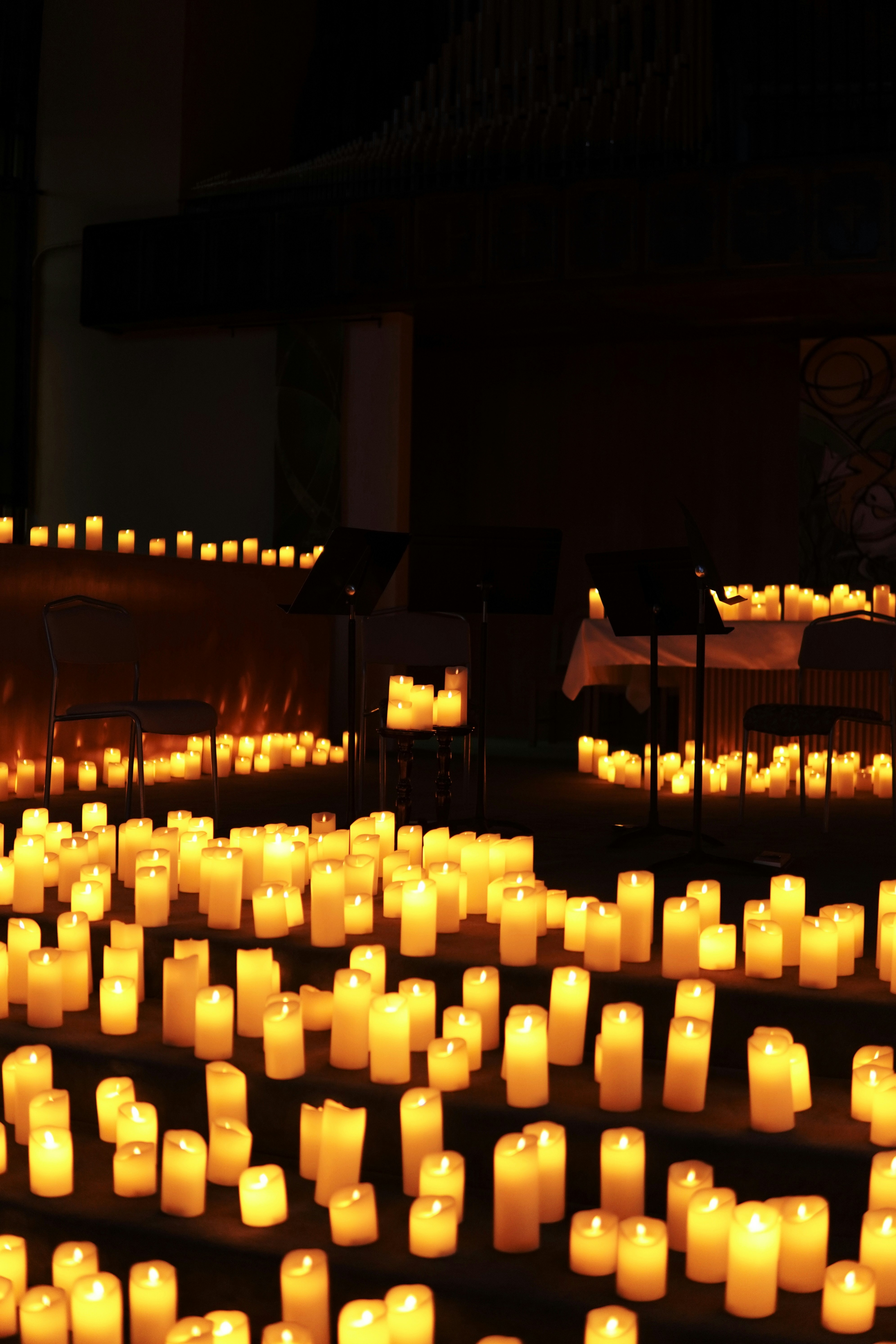 Rows of illuminated white pillar candles arranged on tiered steps in a dim auditorium or church setting. Behind the warm sea of candlelight, music stands and chairs are positioned for a performance, creating a serene atmosphere.