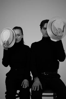 Two people hide behind hats in black turtlenecks.