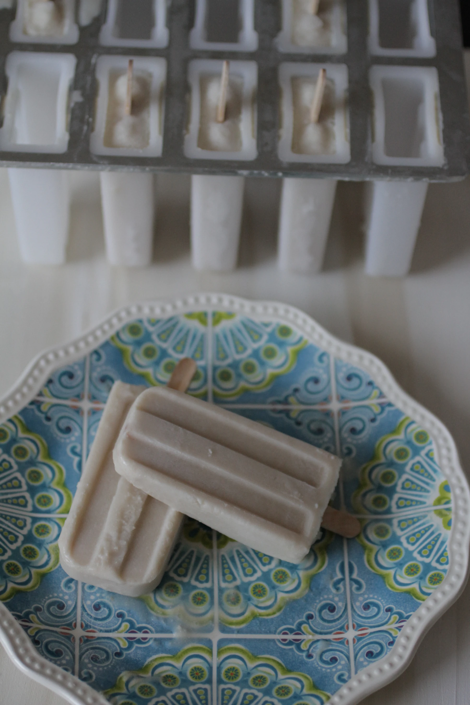 A metal pop mold with wooden sticks set in a creamy base, two unmolded pops on a blue patterned plate in front