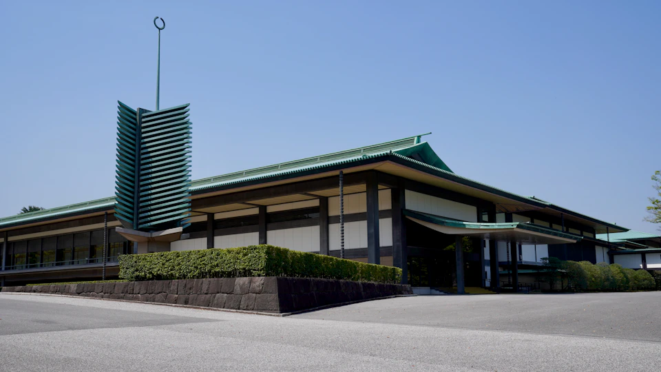 The image shows a large, modern building with a green roof and white walls. The building has a curved facade that adds to its contemporary design. A tall tower stands on top of the structure, reaching towards the sky. The building is situated in an open area surrounded by lush greenery, including trees and bushes.