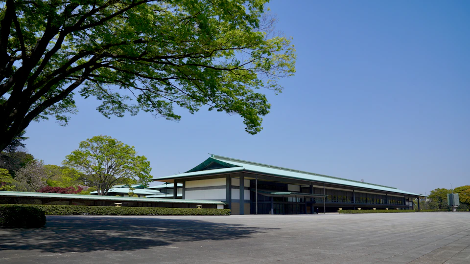 The image shows a large, modern building with a green roof and white walls situated on a street corner. The building is surrounded by lush trees, creating a serene atmosphere. In front of the building, there are several people walking around, adding life to the scene. The perspective of the photo is taken from across the street, providing a comprehensive view of the building's exterior and its surroundings.