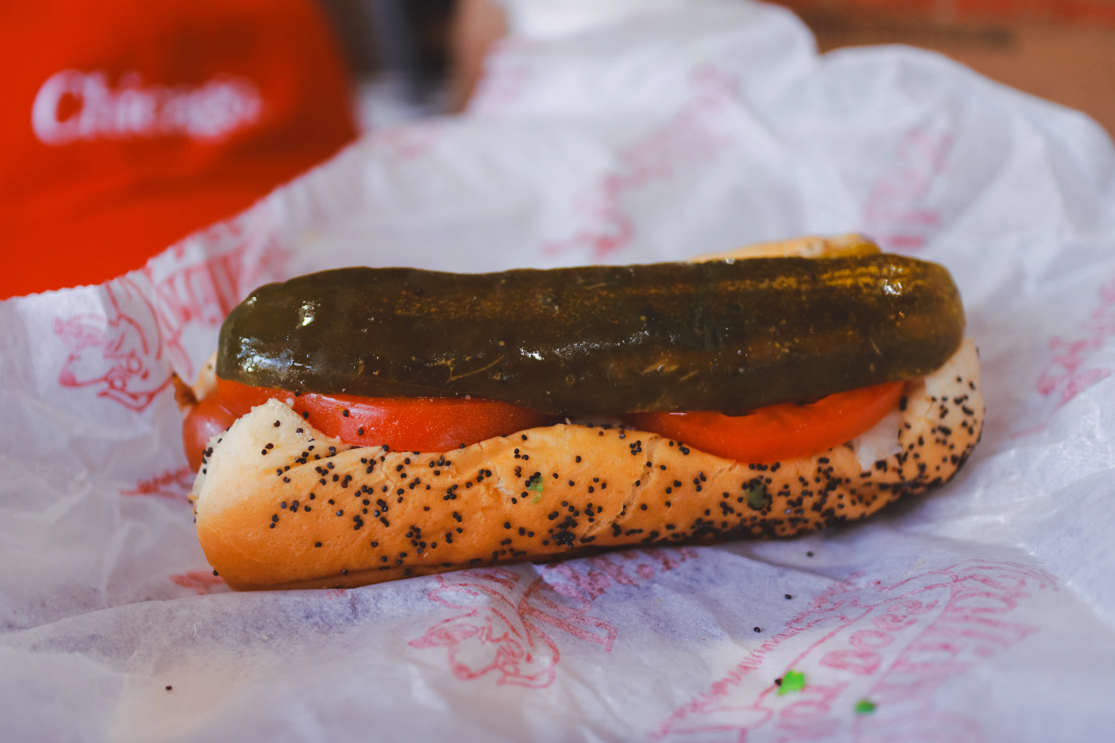 A classic Chicago-style hot dog - poppy-seed bun, tomato wedges and a whole dill pickle spear - resting on its paper wrapper