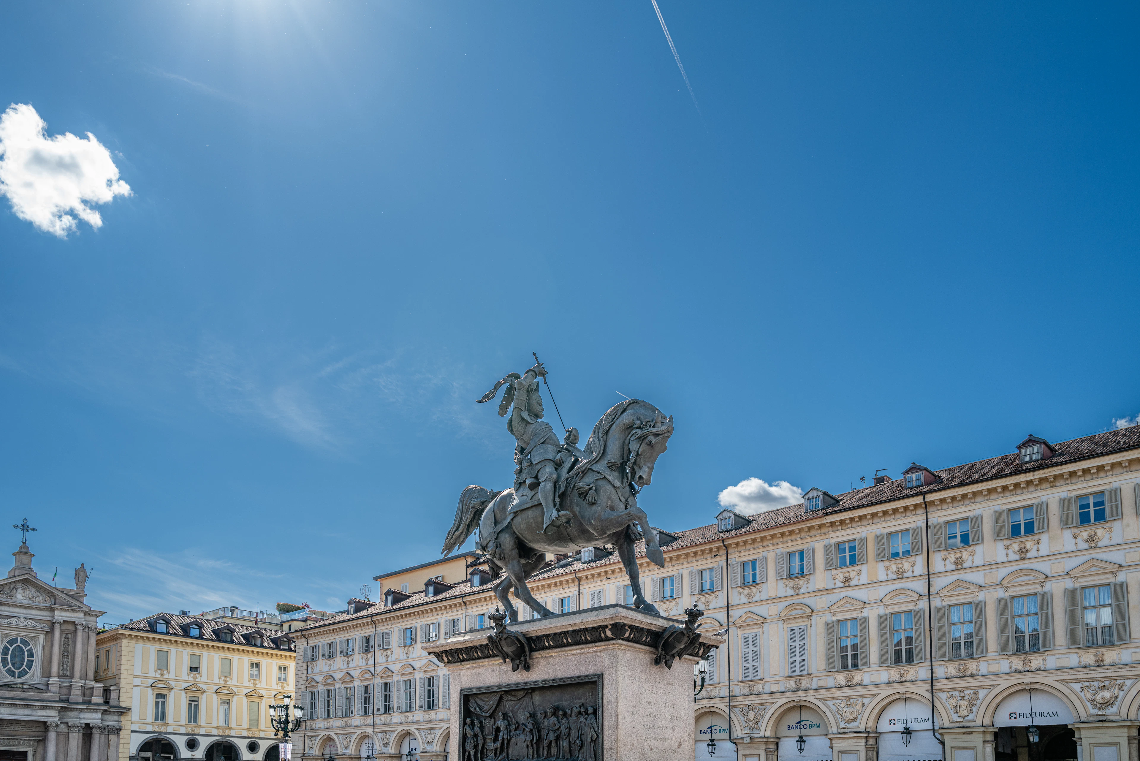 The bronze equestrian statue of Emanuele Filiberto in the middle of Turin's Piazza San Carlo, surrounded by the pale baroque palazzi and arcades of the square under a deep blue sky.