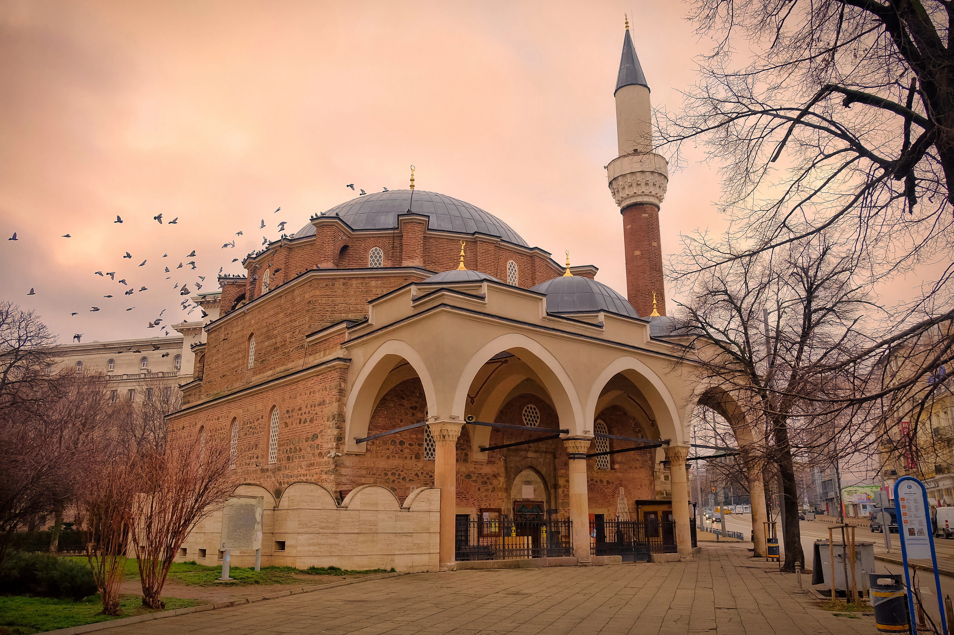 The brick dome, arcaded porch and slim minaret of Sofia's Ottoman-era Banya Bashi Mosque at dawn, pigeons scattering across a pink and apricot sky above bare winter trees.