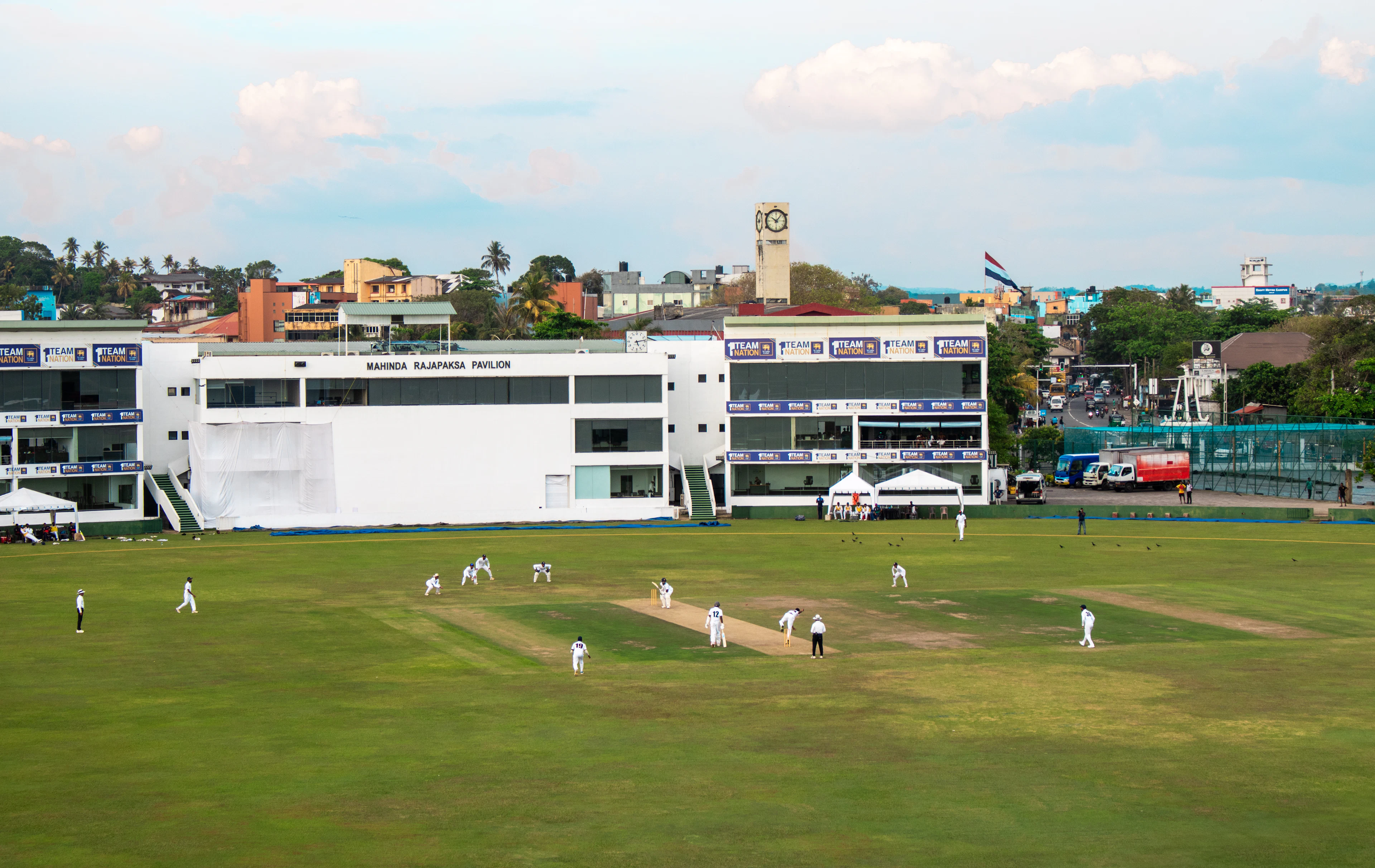 Cricket match in progress with players in whites and a historic clock tower behind the ground