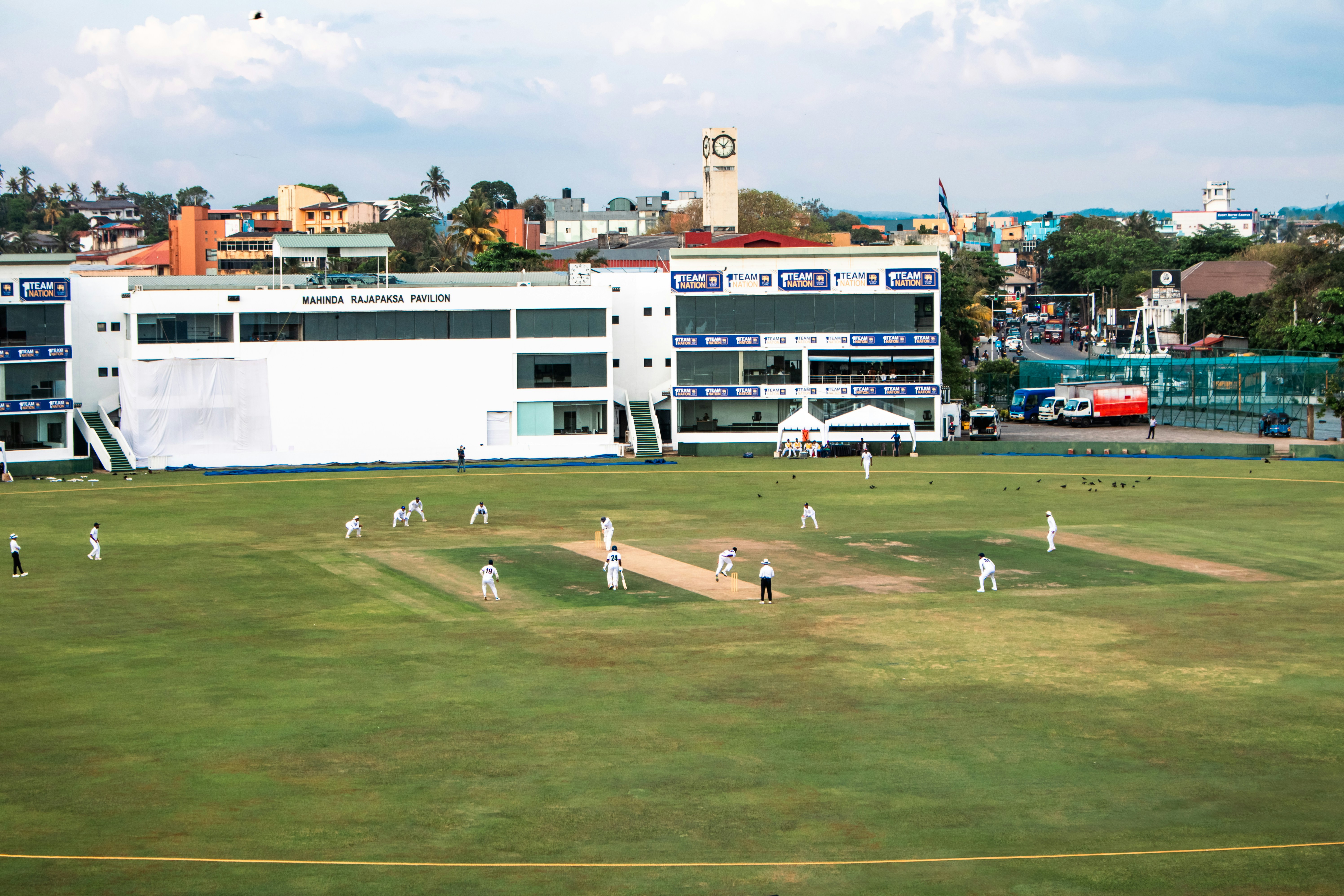 A cricket match unfolds at the Galle International Stadium, Sri Lanka. Players in white dot the lush green field around the central pitch. The Mahinda Rajapaksa Pavilion forms a backdrop, overlooked by Galle's distinctive clock tower and town buildings. Renowned for hosting international and domestic cricket, this ground provides a unique venue for observing Sri Lanka's hugely popular national sport amidst historic and natural surroundings.