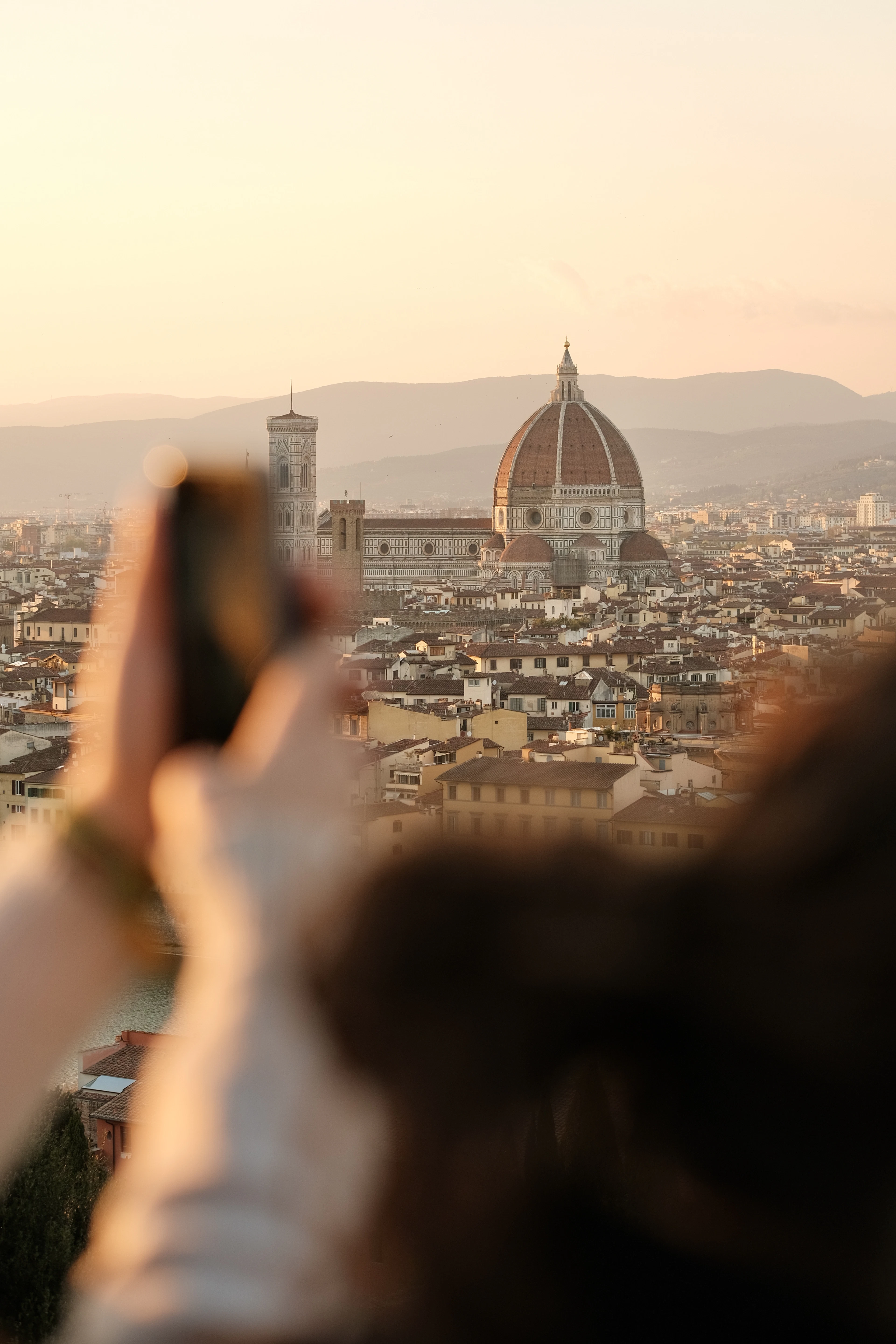 A visitor raising a phone to photograph the Duomo and campanile from a hilltop viewpoint over Florence in soft golden evening light