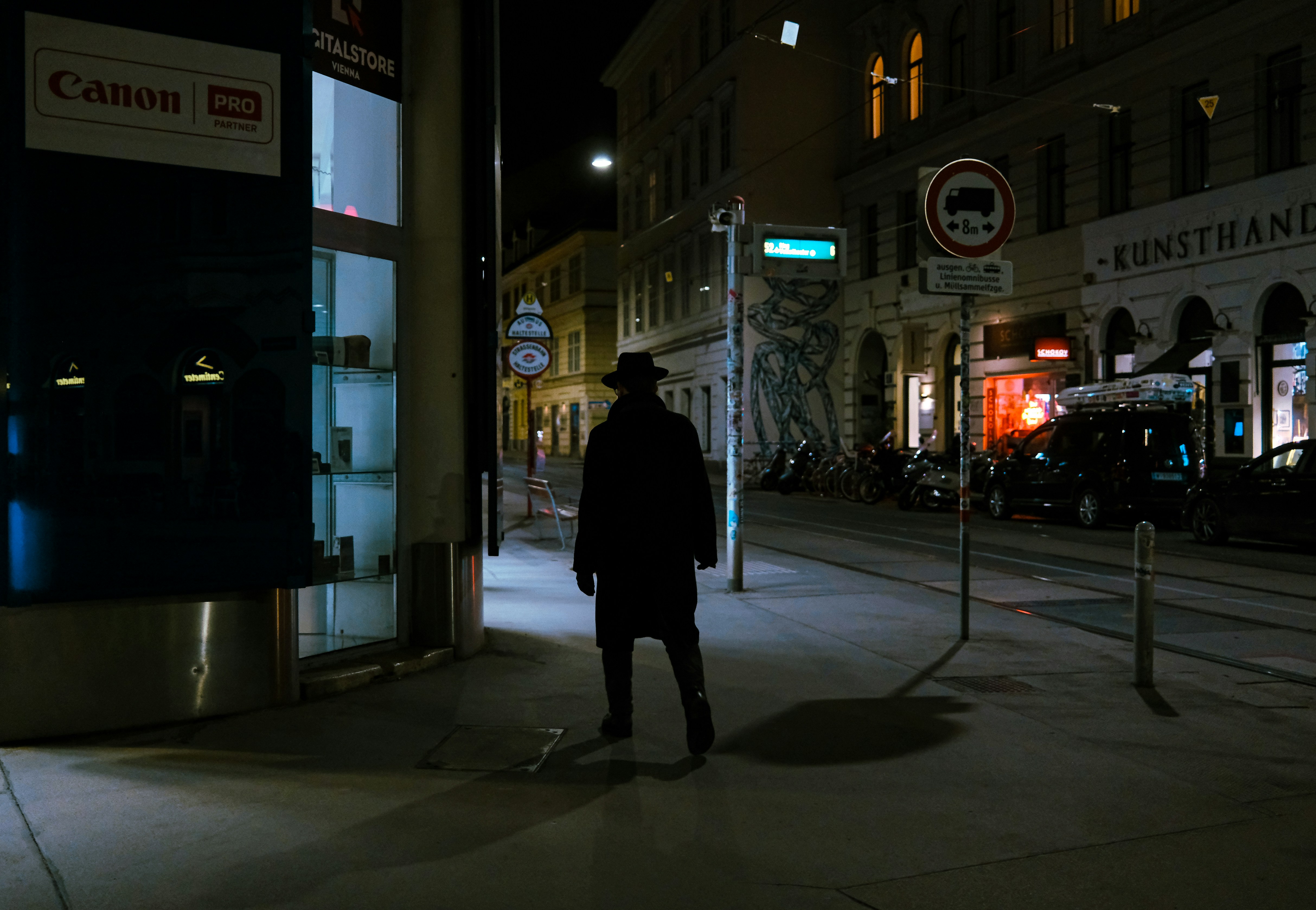 A man in a hat walks down a dark street.