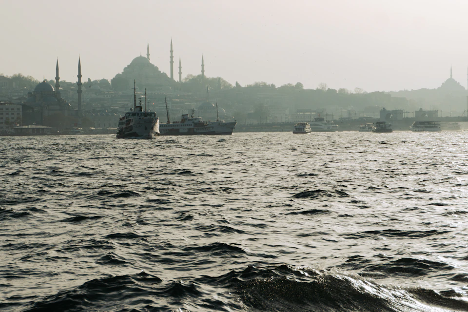 The image depicts a serene harbor scene with several boats floating on the water, including a large ship and smaller boats. The sky above is hazy gray, creating an atmospheric backdrop for the tranquil waters below. In the distance, there are buildings visible along the shoreline, adding depth to the scene.
