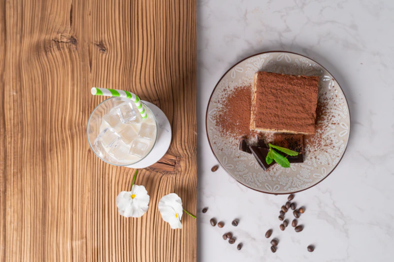 A square of tiramisu dusted with cocoa beside an iced drink and scattered coffee beans