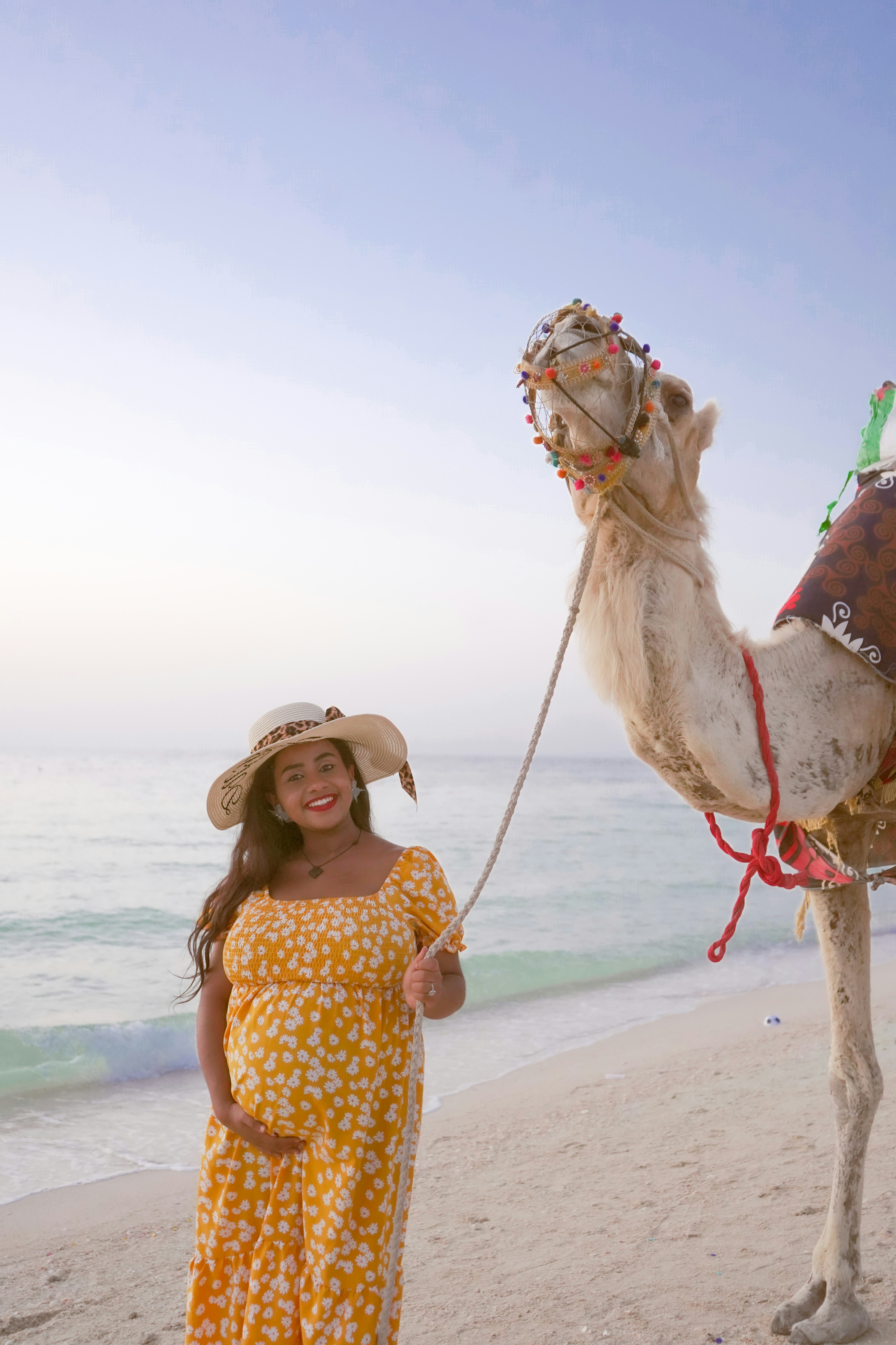 Pregnant woman smiles next to a camel on a beach.