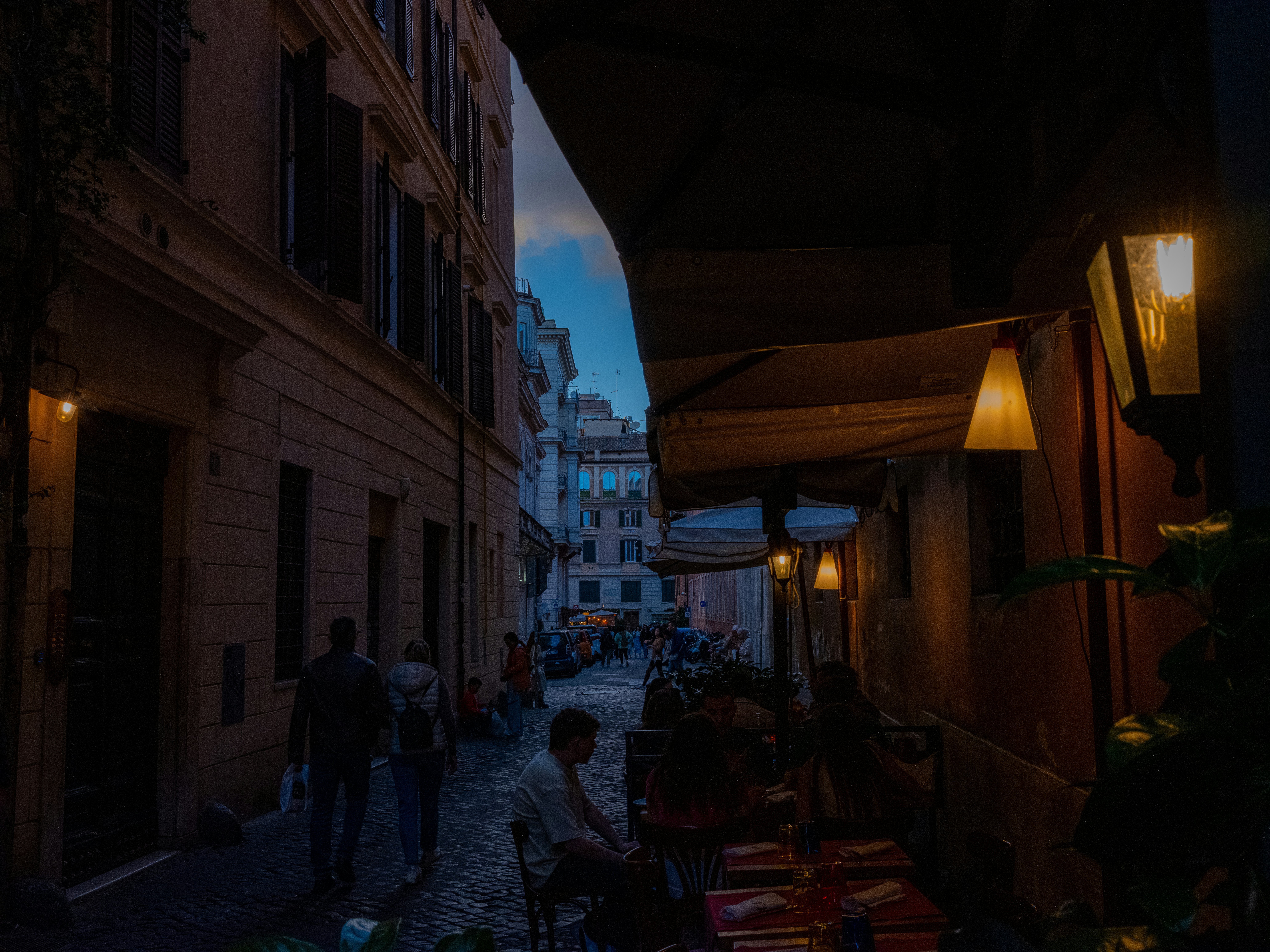 A cozy evening scene in a charming Roman alleyway, where warm lights from a local trattoria invite passersby to enjoy authentic Italian dining under the twilight sky.