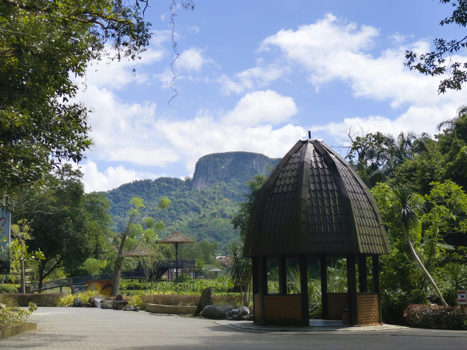A small, dark-roofed gazebo on a path. Behind it is foliage, landscaping, and a few other small structures. In the far background is a huge green hill under a blue sky peppered with clouds.