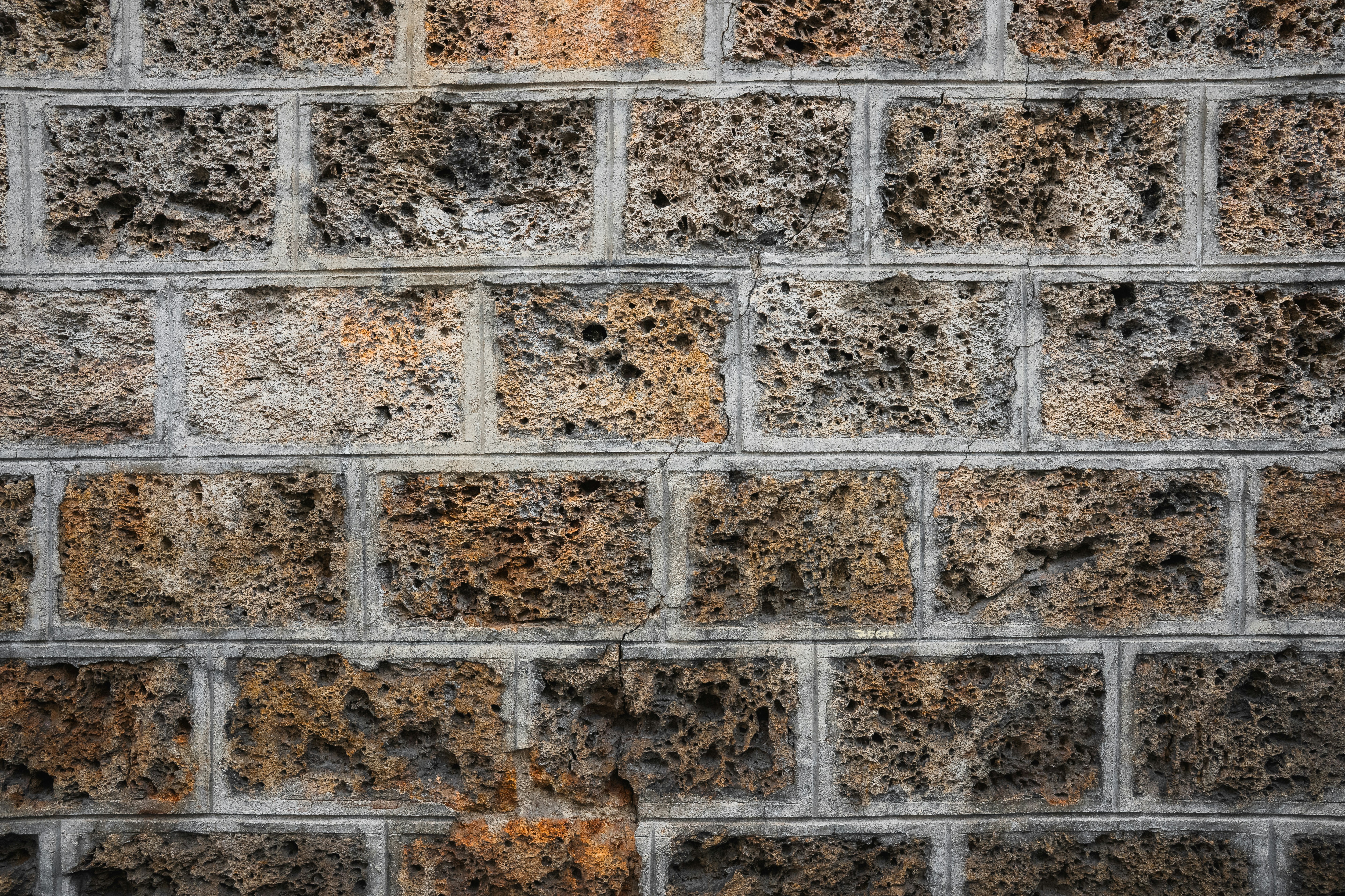 A rugged stone wall made of porous volcanic rock, each brick showcasing a spectrum of earthy tones from deep charcoal to rusty orange. The texture is raw and weathered, evoking a sense of ancient craftsmanship and natural resilience.