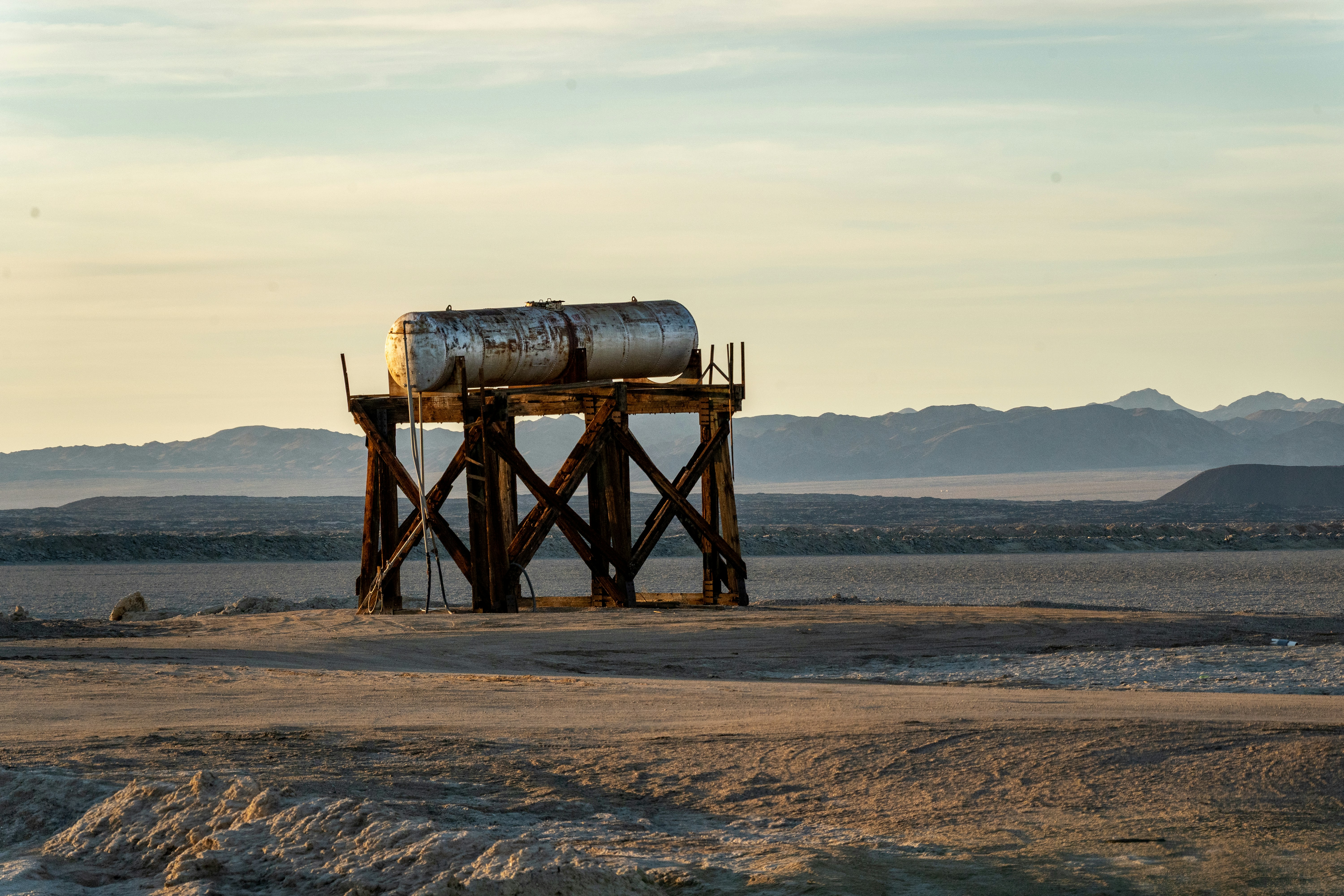 A rusted industrial water tank stands elevated on weathered wooden supports, isolated in a vast desert landscape under soft golden light. The distant mountains and dry terrain evoke a sense of abandonment and endurance in a forgotten corner of the earth.