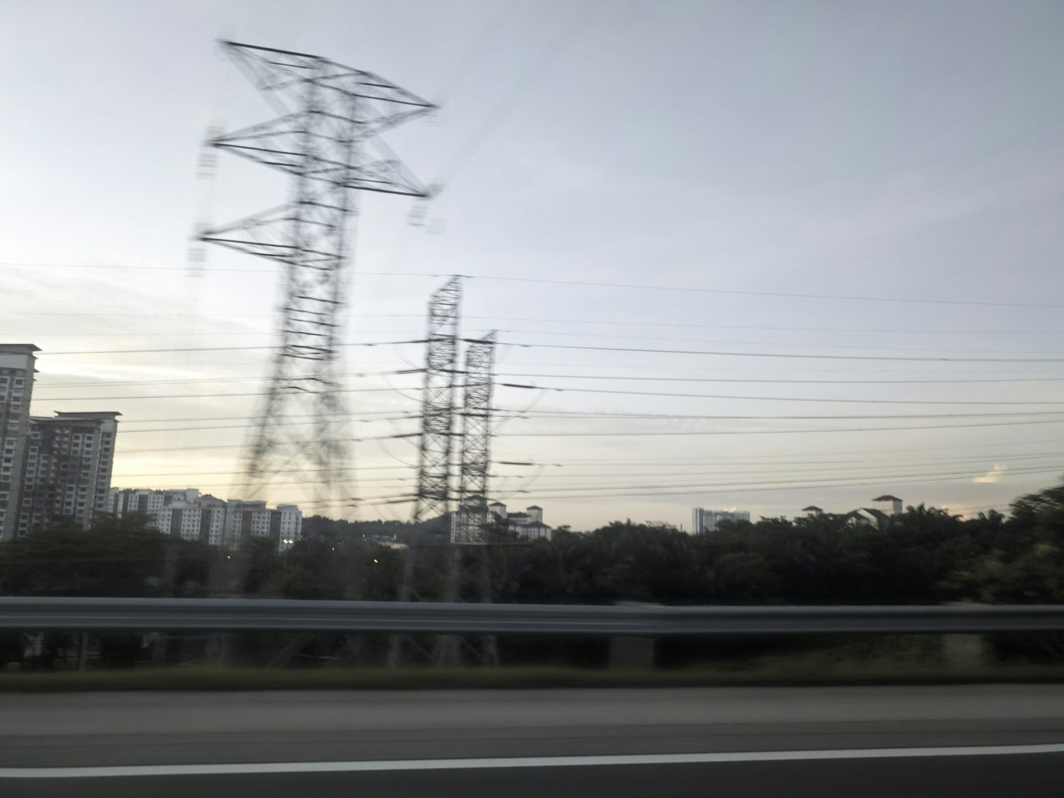 Large electricity pylons and power lines dominate the frame against a cloudy morning sky, with buildings and trees visible in the background, viewed from behind a blurred guardrail.