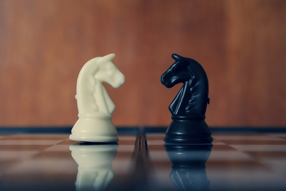 A white knight and a black knight facing each other on a glossy chessboard