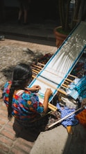 Woman weaves on a loom outdoors in sunlight.