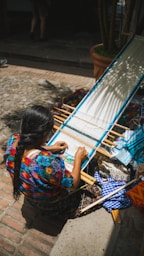 Woman weaves on a loom outdoors in sunlight.