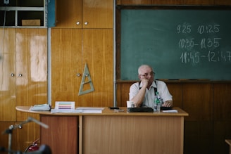 A teacher sits at his desk in a classroom.