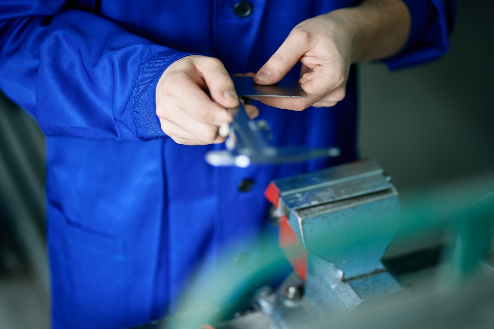 A worker in a blue coat measures a small metal part with calipers over a bench vise.