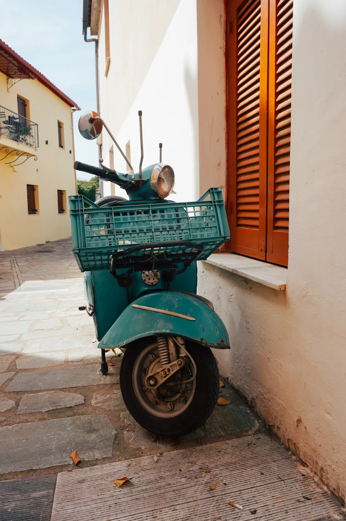 A teal scooter with a crate on the back, parked against a cream wall beside an orange shutter.