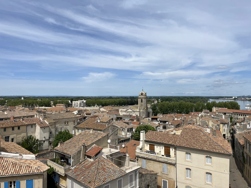 The image presents an aerial view of a quaint town nestled between lush green trees and a serene river. The town is characterized by buildings painted in shades of white, gray, and brown, with some structures featuring red roofs that add a pop of color to the scene. The town's layout is such that it seamlessly blends into the surrounding nature, creating an idyllic atmosphere. In the distance, beyond the town, you can see the river flowing calmly, adding a sense of tranquility to the image.