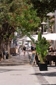 People stroll down a shaded street with cafe seating.
