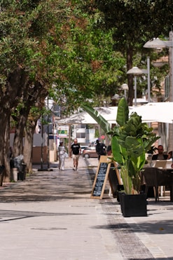 People stroll down a shaded street with cafe seating.
