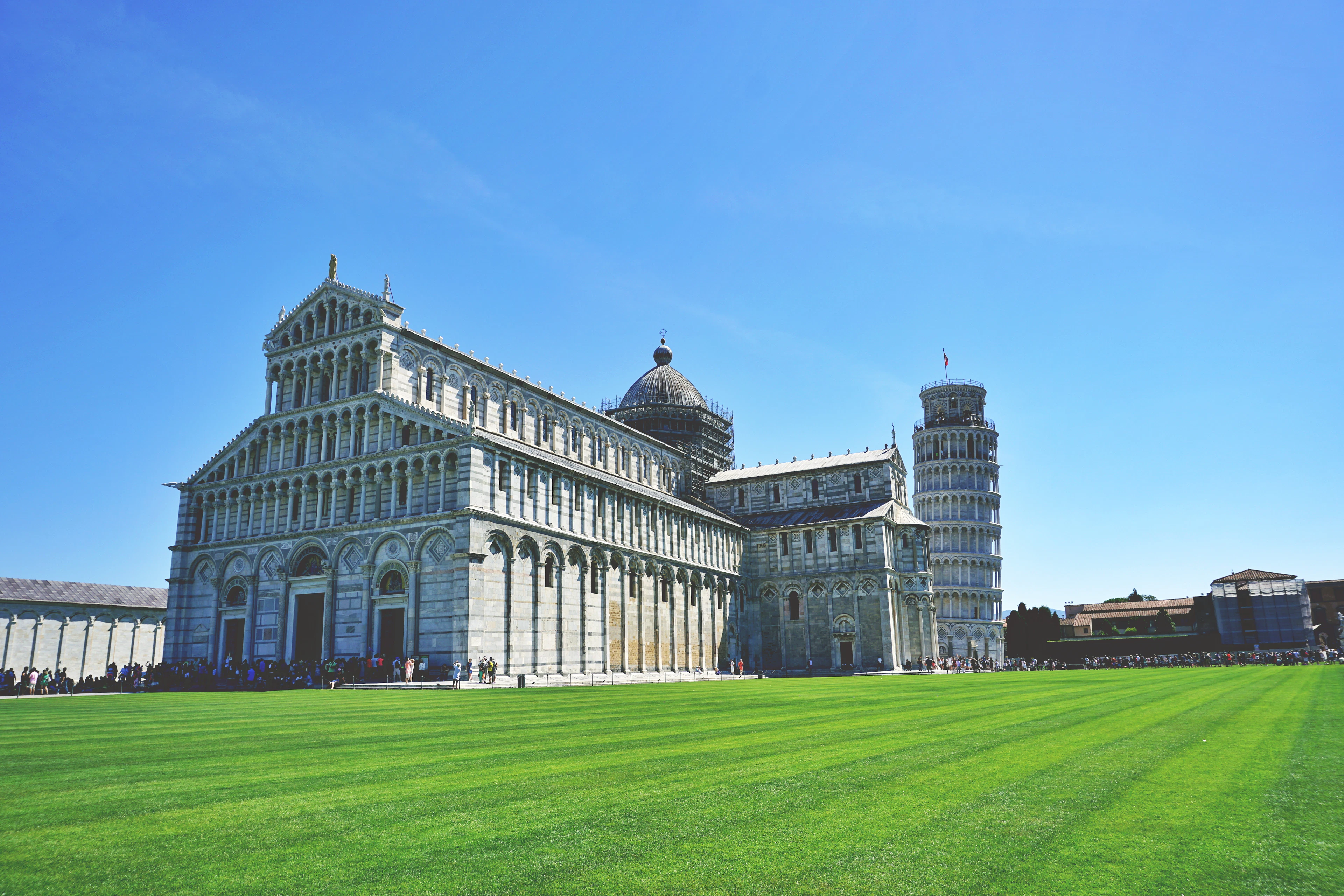 Pisa's cathedral and the Leaning Tower rise from a vivid green lawn under a bright blue sky.