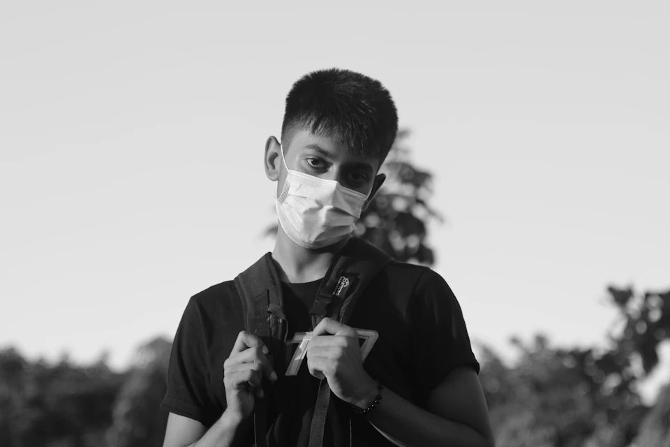 Young man wearing face mask stands against trees, gazing off into distance with backpack