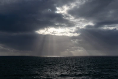 Sunlight pierces through dark, cloudy skies over the sea.