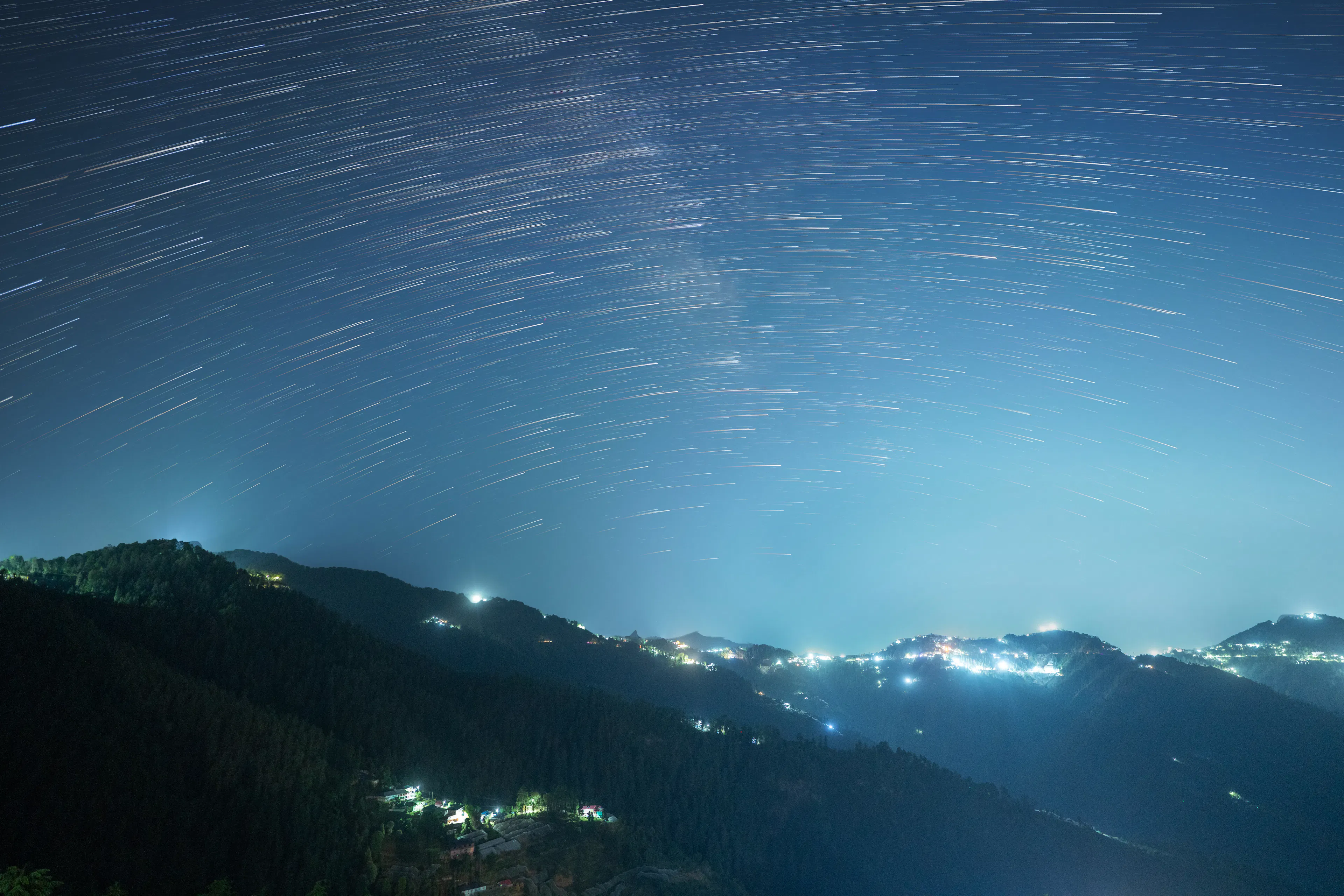 Star trails over a dark mountain ridge with village lights below
