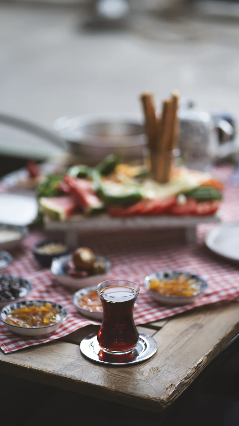 Ahşap masanın ucunda ince belli bardakta çay; arkada kırmızı kareli örtü üzerinde reçel ve zeytin tabaklarıyla kurulmuş uzun kahvaltı sofrası.