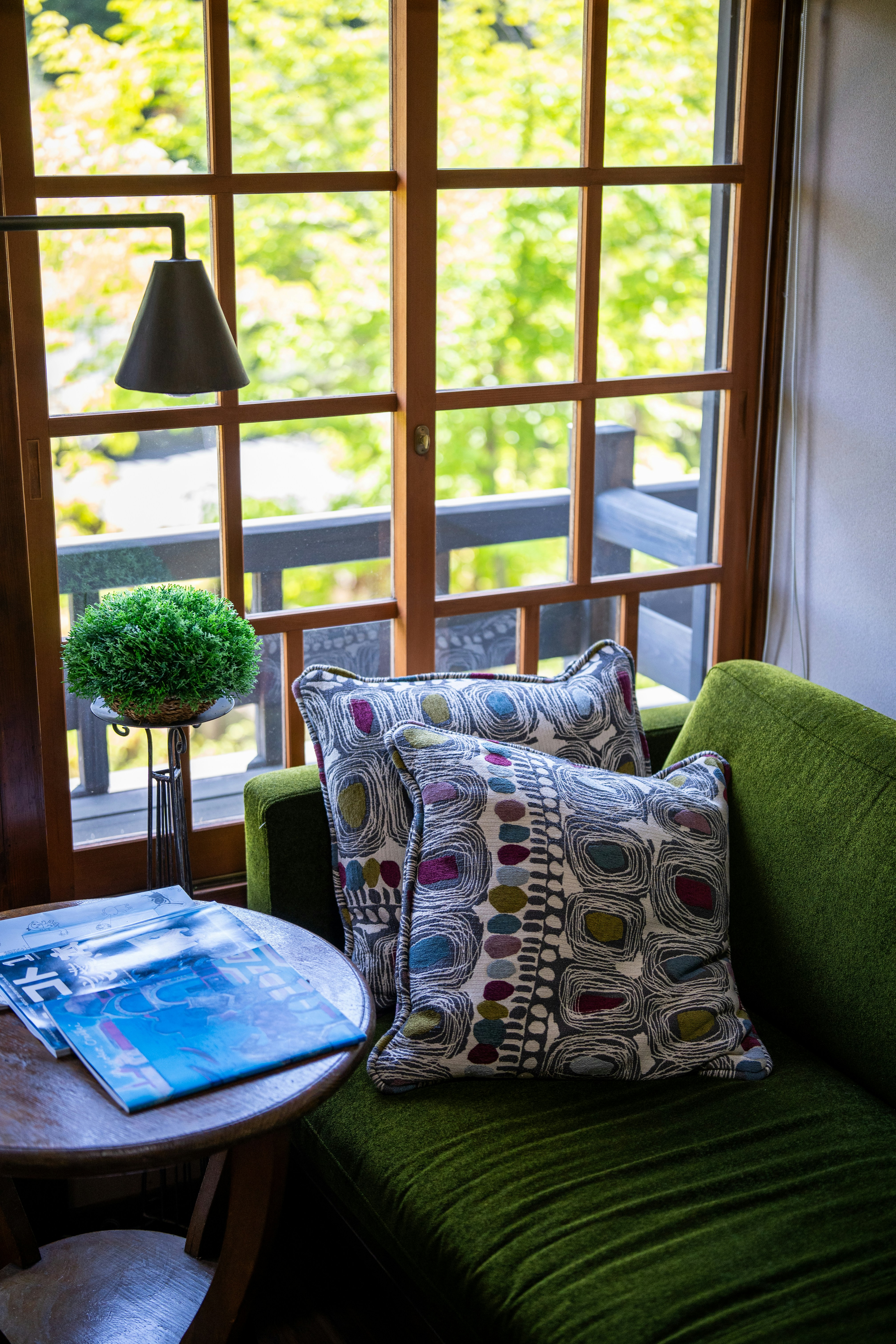 Interior photograph of a cozy living nook featuring a green sofa adorned with patterned cushions, and a round wooden coffee table with a blue magazine. A potted topiary and a black pendant lamp sit by large wooden-framed windows that flood the space with daylight.